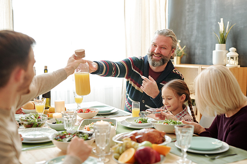 Happy senior man passing bottle of orange juice to his son over served festive table with blonde woman and cute little girl sitting near by