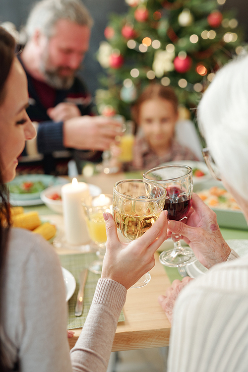 Hands of young woman and her granny clinking with glasses of wine while sitting by served table at Christmas dinner