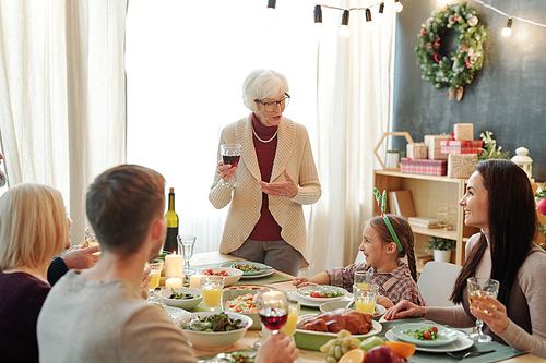 Senior grey-haired woman with glass of red wine pronouncing Thanksgiving toast while standing by served table in front of her family