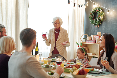 Senior grey-haired female toasting with glass of red wine by served table in front of her family during feestive dinner