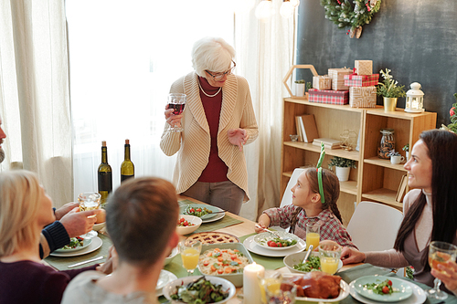 Senior woman with glass of red wine pronouncing festive toast by family dinner while looking at her little great granddaughter