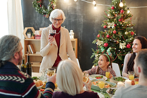Happy senior woman with glass of red wine making Christmas toast while standing by served table in front of big family