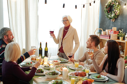 Smiling senior woman holding glass of red wine while toasting by served table in front of her family on Thanksgiving day