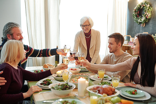 Young and senior members of big family clinking with glasses of wine over served festive table during Thanksgiving dinner