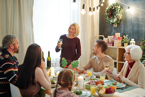 Happy mature female with glass of wine making festive toast by served table while looking at her family during dinner