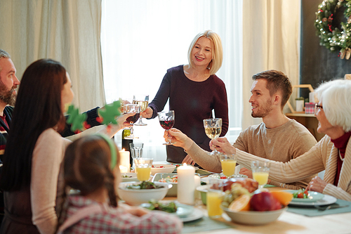 Blonde mature female and her family members clinking with glasses of wine by served table during festive dinner at home