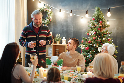 Bearded mature man with glass of wine toasting by served table while standing in front of his family during festive dinner at home