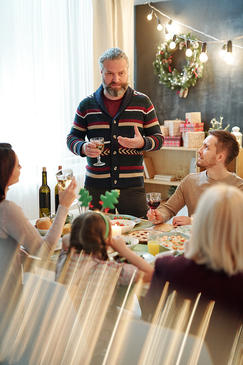 Bearded man with drink pronouncing Christmas toast in front of his family by served table while gathering together at home