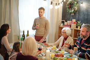 Young man with glass of wine making toast by served table in front of his family during festive dinner at home