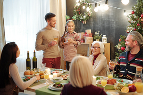 Happy young man with glass of wine and his cute little daughter standing in front of rest of the family at Christmas dinner