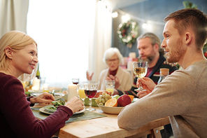 Young man with glass of wine offering festive toast to his happy mother sitting next to him and eating fresh salad