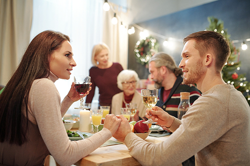 Young affectionate couple with glasses of wine making festive toast by served table on background of big family by dinner