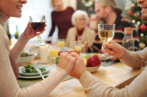 Hands of young affectionate couple with glasses of wine making festive toast on background of big family by dinner