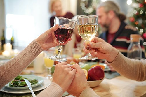 Hands of young affectionate couple clinking with glasses of wine after making festive toast by served table at Christmas dinner