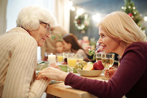 Senior woman in eyeglasses and her blonde daughter chatting by served festive table during family dinner