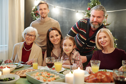 Big family of six looking at you while sitting by served table in front of camera and enjoying festive dinner at home