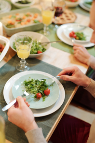 Hands of young man holding knife and spoon over plate with fresh green salad and tomates during family dinner