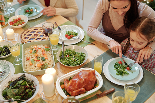 Young mother cutting tomato on plate of her little daughter while both sitting by served festive table and having dinner