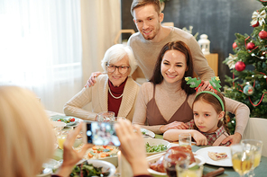Cheerful affectionate family sitting by served festive table and looking at smartphone camera in hands of mature woman