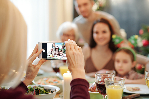Hands of mature female holding smartphone while taking photo of young couple, their little daughter and granny by dinner