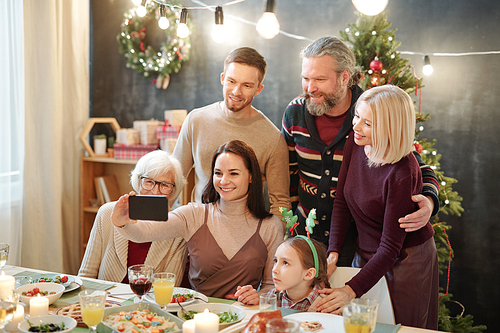 Cheerful affectionate family of six looking at smartphone camera while making selfie by served festive table at home