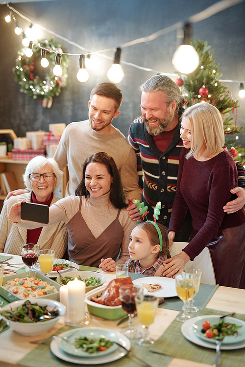 Joyful family of six looking at smartphone camera held by young woman while making selfie by served festive table at home