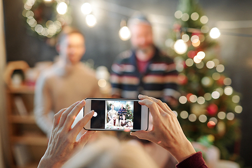 Hands of female holding smartphone with photo of happy affectionate family on its screen while taking photo by festive table