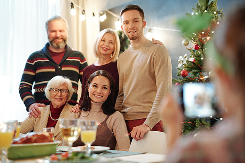 Happy family posing for smartphone camera by served table in front of little girl taking photo of them at Christmas dinner