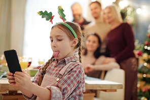 Cute little girl with Christmas headband on head showing tongue while making selfie on background of her family