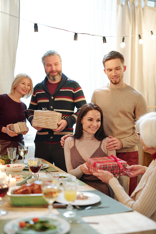 Young affectionate couple giving Christmas present in giftbox to granny sitting by served festive table after dinner