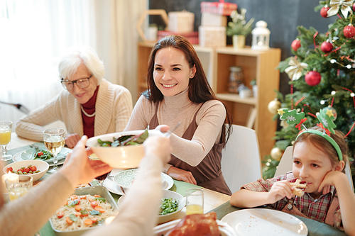 Happy young woman taking bowl with homemade salad from hands of her husband over served festive table