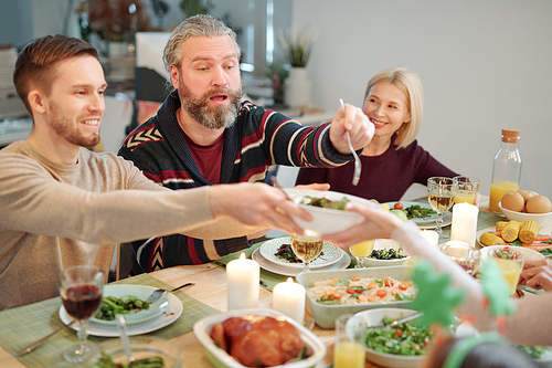 Bearded man with fork going to take some salad from bowl held by his young son over served table during family dinner