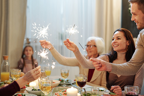 Cheerful young and mature adults holding sparkling bengal lights over served festive table during Christmas dinner