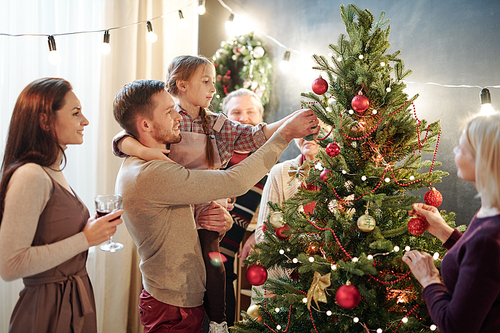 Young man with his little daughter decorating Christmas tree at home among other family members while preparing for holiday