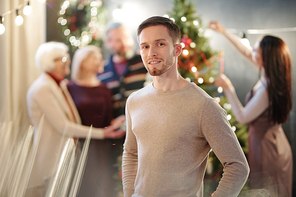 Happy young man in beige pullover standing in front of camera on background of women decorating firtree before xmas