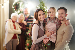 Happy young family of three standing in front of camera on background of mature adults by Christmas tree