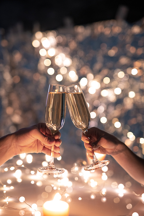 Hands of young couple clinking with flutes of champagne on background of Christmas lights during celebration