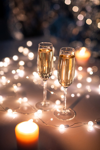 Two crystal flutes of sparkling champagne on table surrounded by lit garlands and burning candles in festive environment