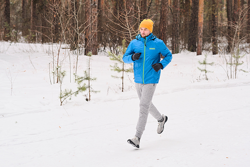 Young athlete in warm hat running along snowy path in winter forest while training alone