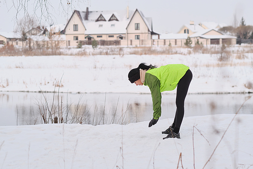 Young brunette woman in green jacket standing at lake and doing exercise before running