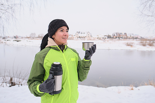 Happy young woman in warm gloves standing at river shore and drinking hot tea during hiking