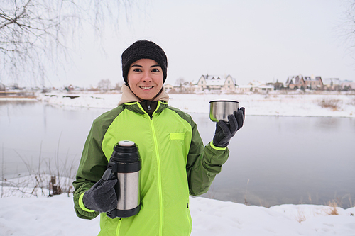 Portrait of positive young woman in green jacket holding thermos and drinking hot tea at winter lake