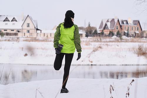Rear view of brunette woman doing quad stretch exercise while stretching leg at winter river coast