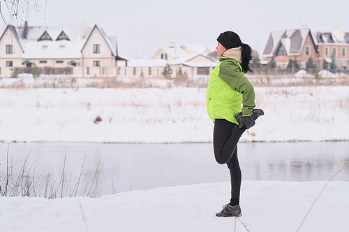 Side view of young woman with black hair stretching leg while preparing for running in winter park