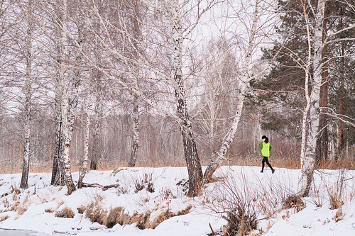 Active woman in green jacket running in winter forest with naked trees