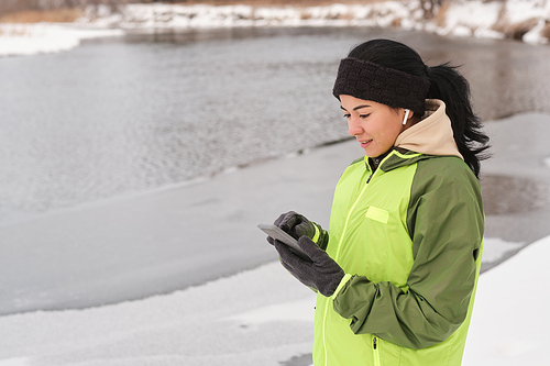 Smiling pretty girl in headband standing on winter shore and texting message on phone