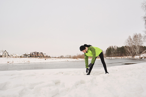 Young woman in green jacket standing t winter lake and bending over while doing warm-up exercise