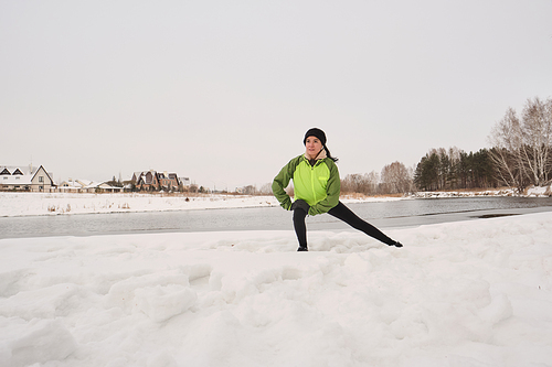 Young athletic woman in jacket performing lunge aside on snow while training on winter shore