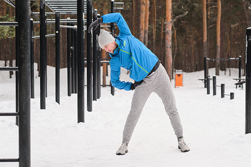 Sporty young man in hat standing at workout area in winter and doing warm-up exercise