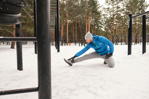 Young man in blue jacket sitting on one knee and stretching leg in crouch position while training on winter workout area